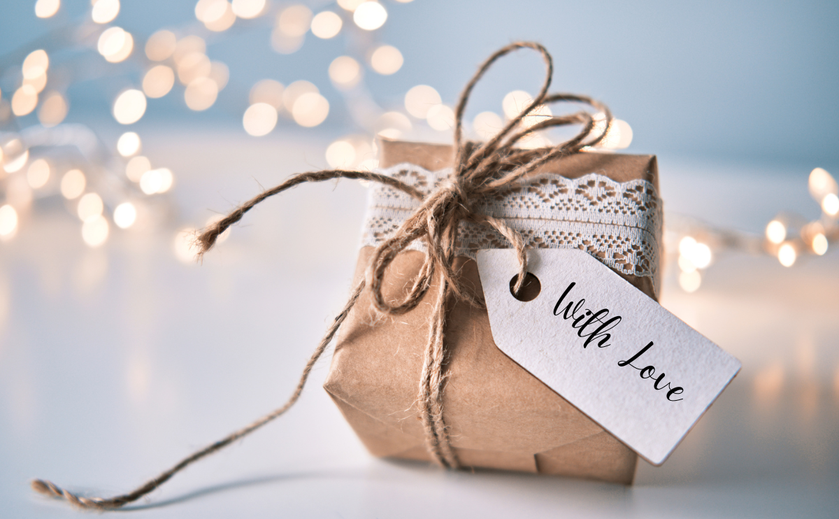 Unique Holiday Gift Ideas, image has a wrapped gift, with a tag that says with love in front of a light blue background with fairy lights.