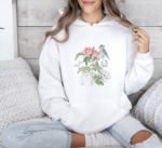 Cottagecore Botanical Bird Hoodie in White, worn by a woman on a soft sofa.